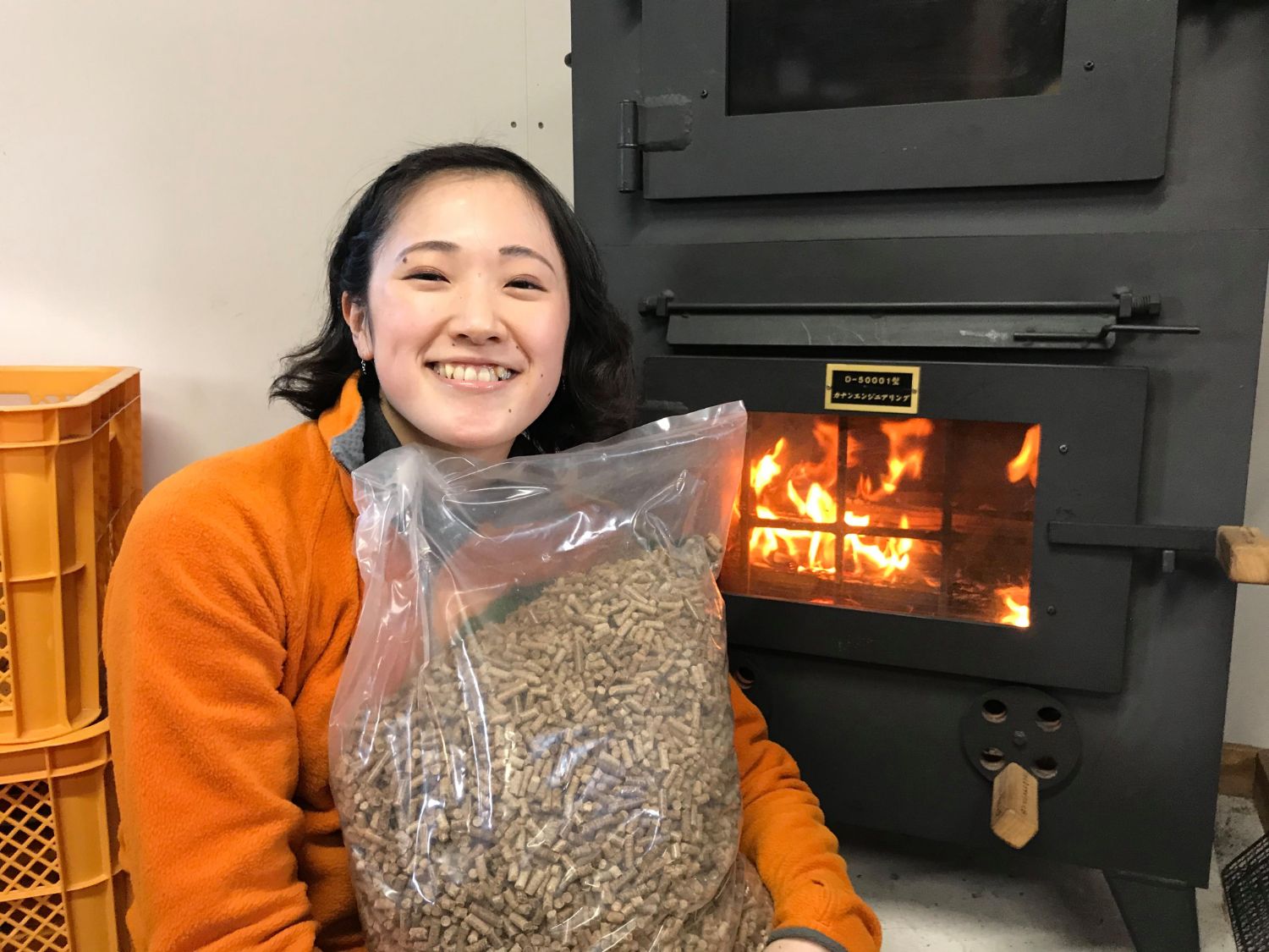 薪ストーブの前でオレンジ色のフリースを着た女性が、木質ペレットの入った大きなビニール袋を抱えて笑っている写真