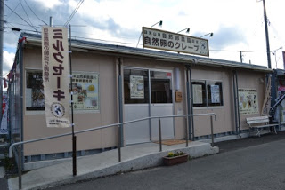 看板に「自然卵のクレープ」と書かれており、入り口にスロープが設置され、ミルクセーキと書かれたのぼり旗が立っている、自然卵農園直営の自然卵のクレープ モアイ店の外観写真