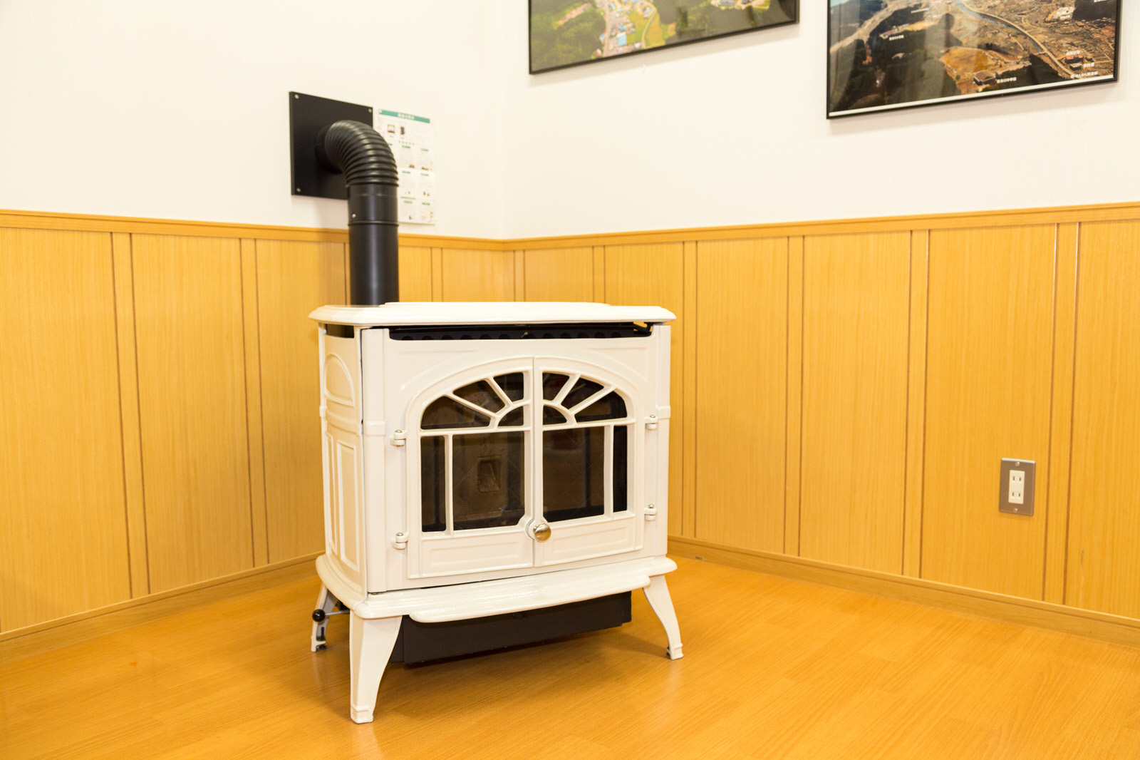 航空写真が額に入って飾られている木の板張りの室内の壁に黒い排気管が接続されており、手前に白いペレットストーブが設置されている写真