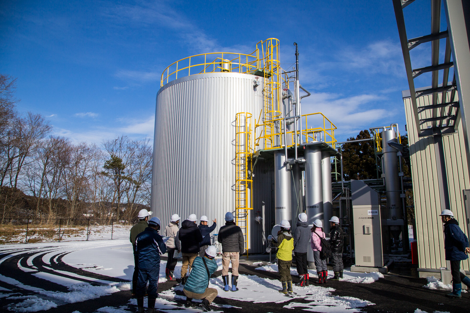 雪がうっすら積もった地面の上で、ヘルメットをかぶった複数の人々が大型の金属製タンクや配管設備が設置されたバイオガス関連施設を見学している様子の写真