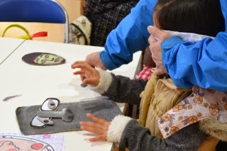 子どもが目隠しをしてモアイの顔のパーツを貼る福笑いに挑戦しており、大人がサポートしている正月遊びの写真