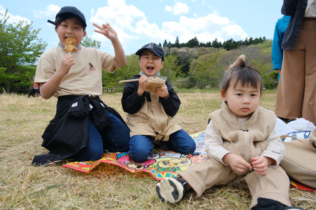 芝生の上にレジャーシートを敷き、左の男の子は焼き鳥を食べながらピースサインをし、中央の男の子は焼きそばを食べて嬉しそうに笑い、右の幼い女の子は静かに座っている、イベントを楽しむ子どもたちの写真