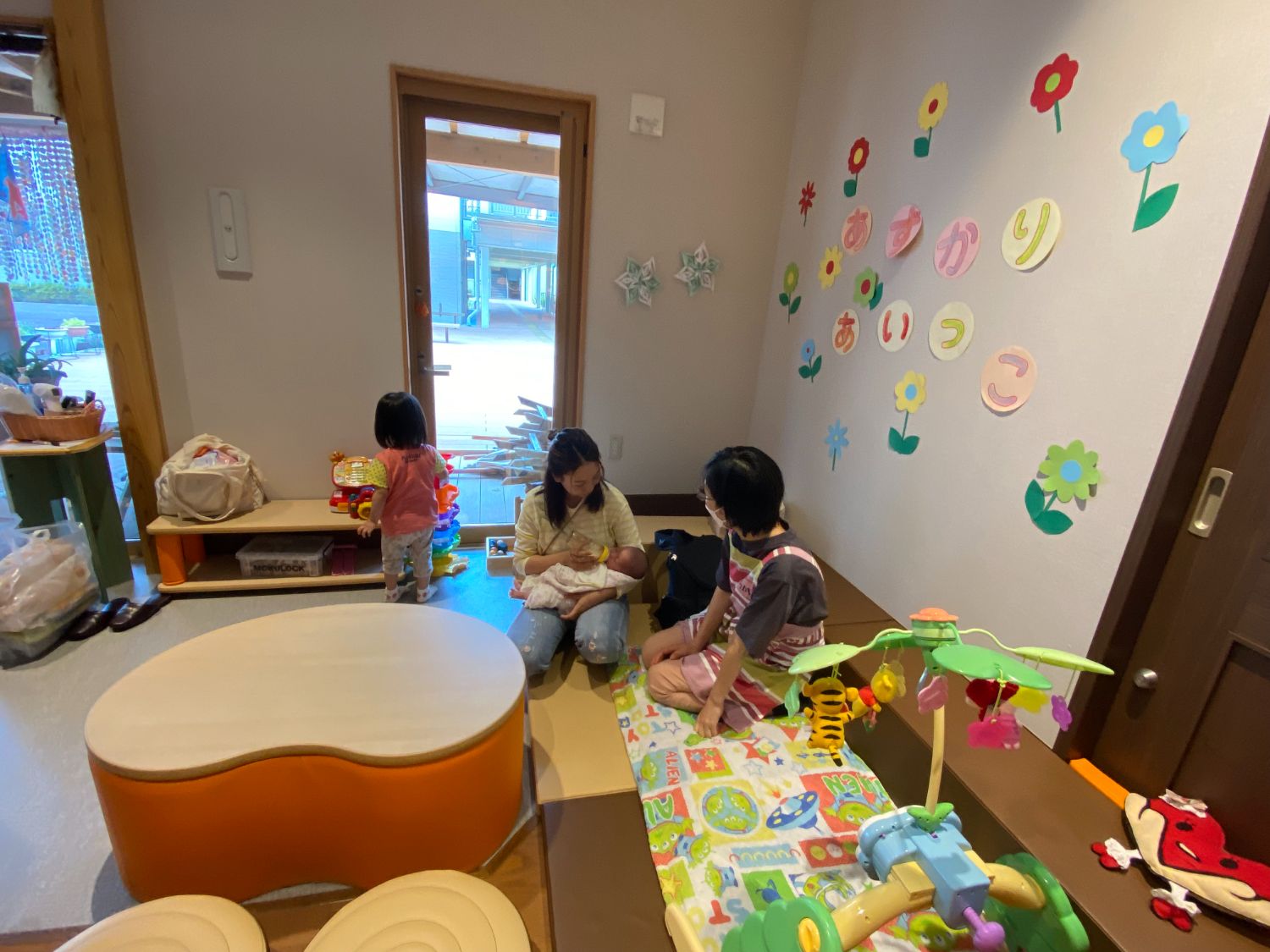床に座ったお母さんが赤ちゃんを抱き、その隣でもう一人の保育士が話しかけており、別の幼児が近くの玩具台で遊んでいる様子の写真