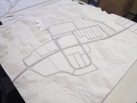 白い地形模型の上に灰色の道路と区画が描かれ、街の基本設計を表している段階の模型の写真