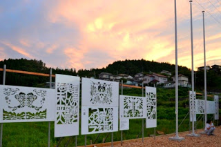 夕焼けの空を背景に、地元の人たちが制作した切り絵パネルが屋外のネットフェンスに展示されている写真