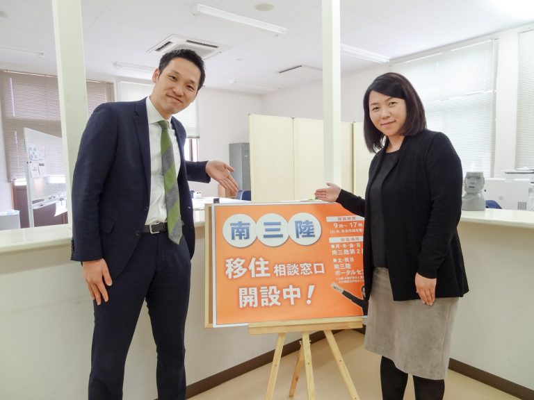 スーツ姿の男女が「南三陸 移住相談窓口 開設中!」と書かれたオレンジ色の看板を紹介するように指さしている写真