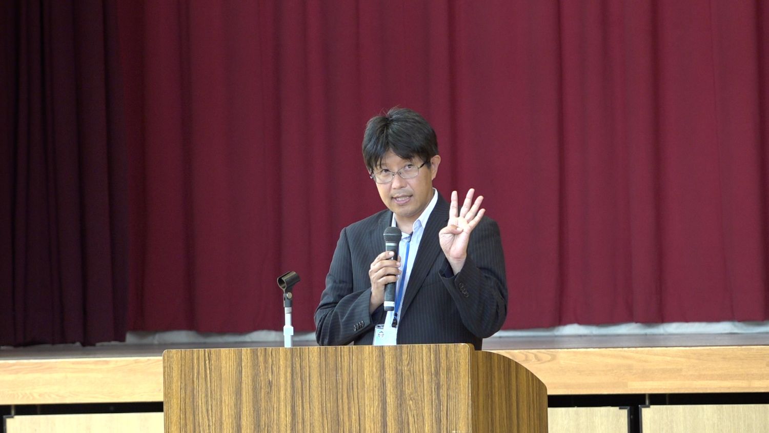 スーツ姿の男性が体育館の演台でマイクを持って話しながら片手で4本の指を立てて説明をしてしている様子の写真
