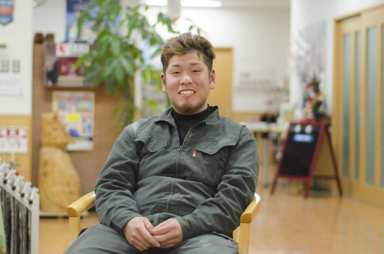 室内の椅子に座った、カーキ色の作業服を着た笑顔の千葉さんの写真