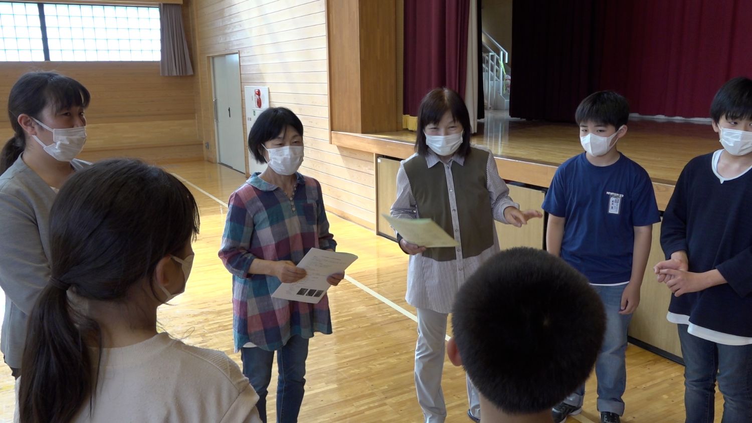 体育館のステージ前で、数人の女性がプリントを手にしながら小学生たちと向き合って話しており、みなマスクを着用していて、地域の人々が子どもたちに小太鼓のコツを子どもたちに伝授する様子の写真