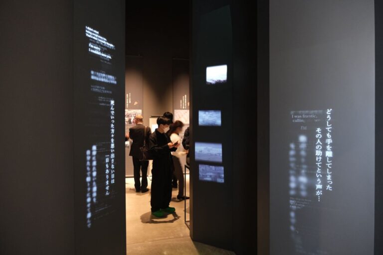 左右の黒い壁に文章がや映像が投影された、狭い入り口の奥の展示スペースで数人の来場者が展示を見ている様子を写した写真