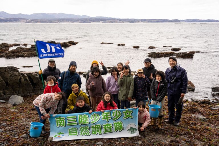 岩場の海岸に立つ「南三陸少年少女自然調査隊」のメンバーたちが、団体名の書かれた横断幕と青い旗を掲げて並んでいる集合写真