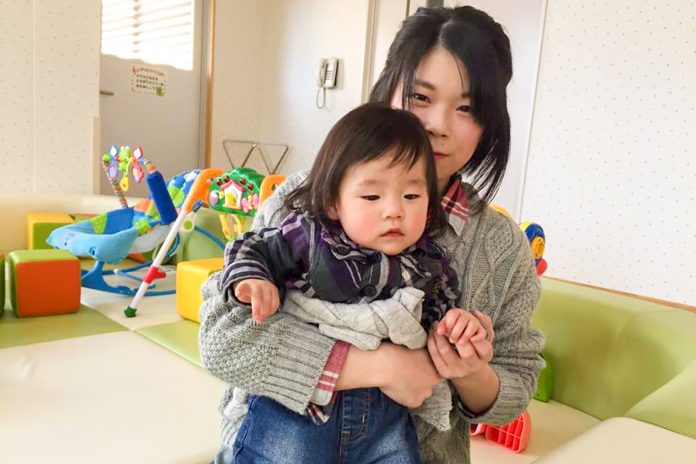 クッションマットが敷かれ、幼児用のおもちゃなどが置かれたスペースに座り、小さな女の子を抱いている佐々木佑委さんの写真