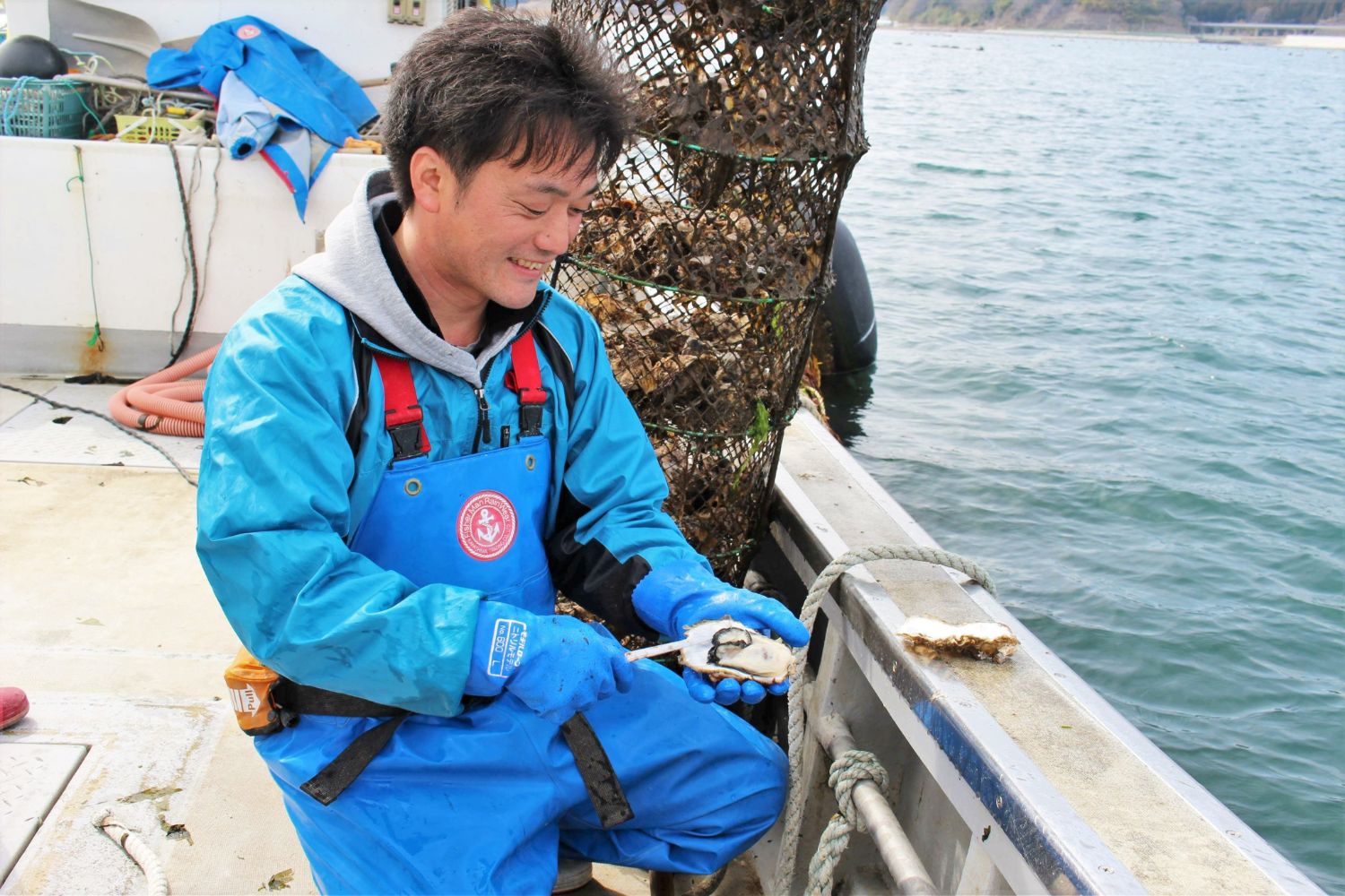 青い作業服を着た後藤新太郎さんが船上で開いた牡蠣を持っている写真