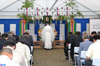 神主が祭壇の前に立って儀式を執り行い、出席者たちが頭を下げて参列しているテント内の様子を写した写真