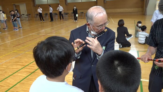 スーツ姿の男性が体育館で子どもたちに篠笛の吹き方を教えており、子どもたちは熱心に聞いている様子の写真