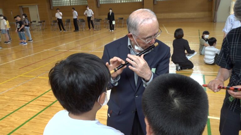 体育館の中でスーツ姿の男性が横笛の吹き方を熱心に子どもたちに教えており、周囲では他の子どもや大人が活動している様子の写真