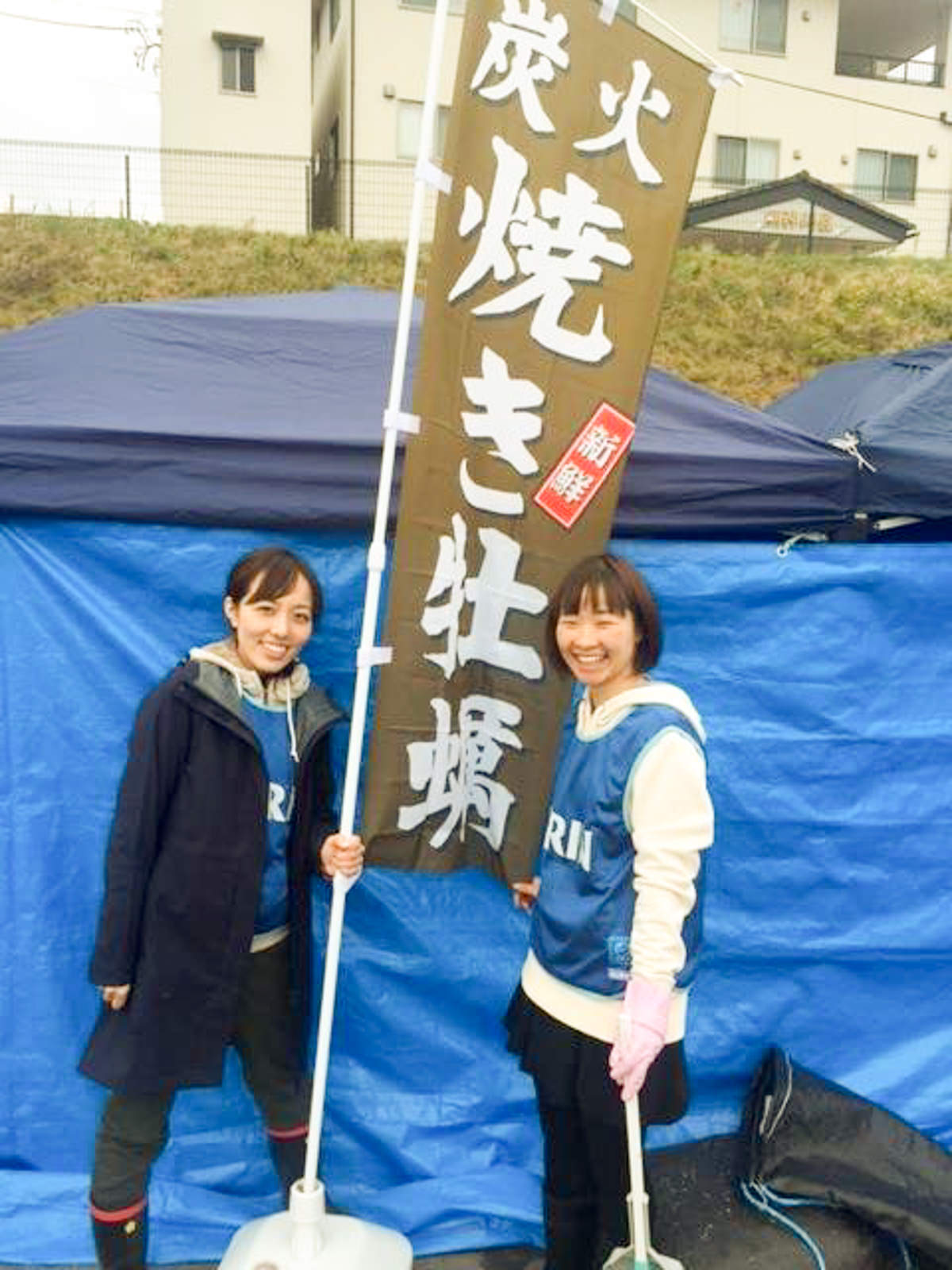 ブルーシートで壁が作られたテントの前で、「炭火焼き牡蠣」と書かれたのぼりの左右に、2人の女性が笑顔で立っている写真