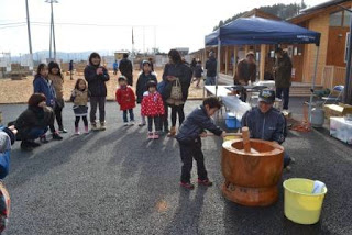 屋外で子どもが大人の指導を受けながら大きな杵で餅つきを体験している様子を写した写真