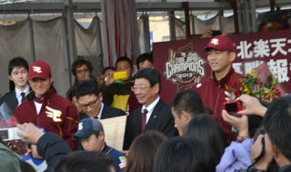 ステージ上で、牧田明久外野手と斎藤隆投手が並び、中央に立つスーツ姿の佐藤町長と関係者たちが、多くの観客に囲まれながら笑顔を見せている写真