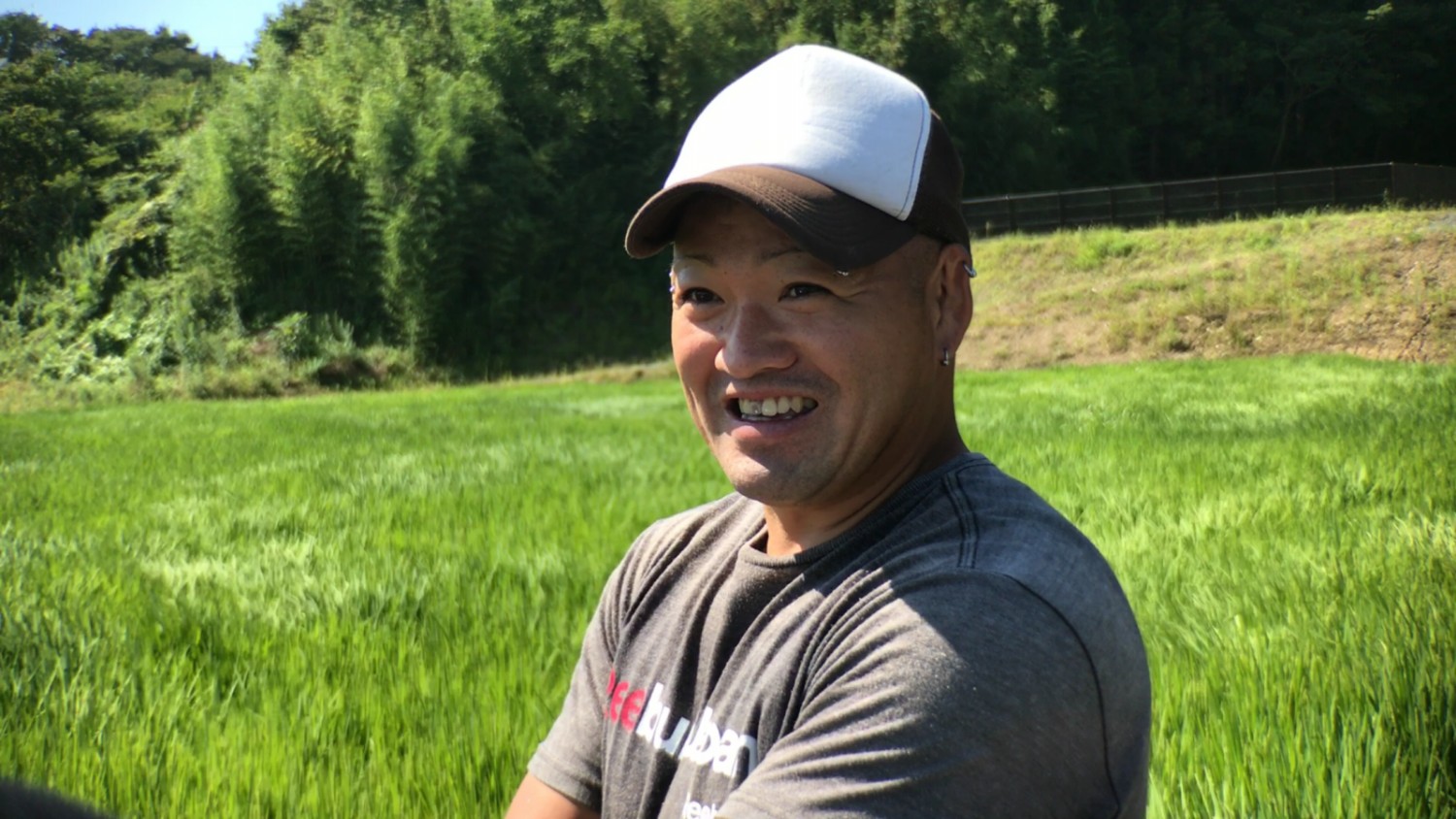 日差しの強い晴れた日、青々とした葉が伸びた畑の前に立ち笑顔で話している渡部さんの写真