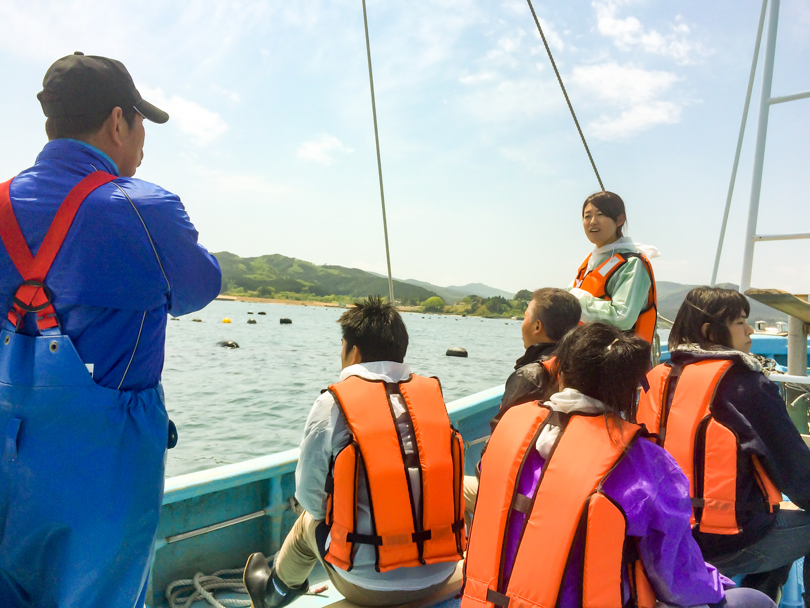 海と山を背景に、ライフジャケットを着た複数の男女の参加者が漁船の上で、立っている青い作業着の漁師と参加者の女性の話を聞いているツアーの様子の写真
