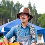 森林が広がり、青いテントが設置された野外イベントの会場で、青いシャツにオーバーオール姿の男性がギターを肩にかけて笑顔で立っている様子をアップで撮影した写真