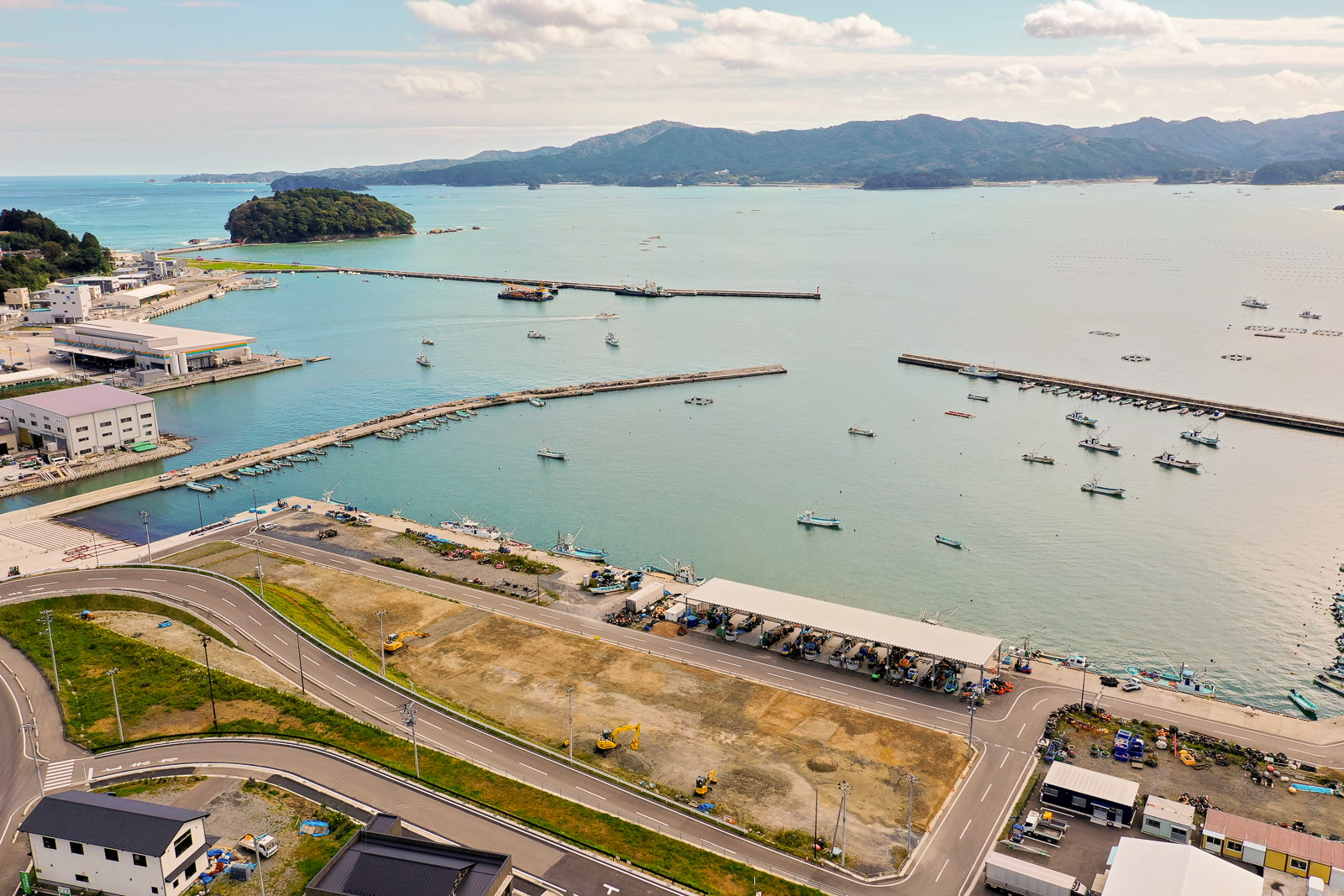 広く青い海が広がり、漁船が停泊する港の周囲にはコンクリート製の防波堤が伸び、湾岸には倉庫や作業場などが並び、駐車場や数台のショベルカーなどの重機も点在している写真