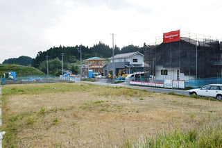 草地の手前に空き地が広がり、その奥では複数の住宅が建設中で足場とシートがかけられている写真