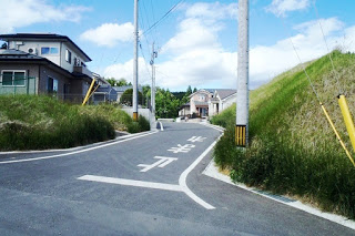 草の生えた法面の間を抜ける道路の先に住宅が見え、路面には「止まれ」と白く書かれている住宅街の写真