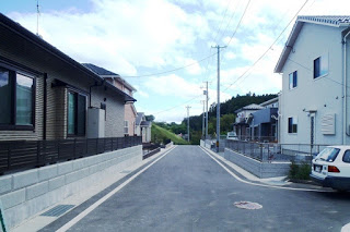 両側に新しい住宅が整然と建ち並び、真っすぐ続く舗装道路の先に緑のある丘が見える静かな町並みの写真