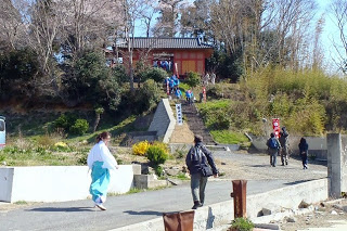 階段を上った先にある赤い鳥居と小さな神社があり、その参道を複数の人々が歩いており、手前には白と水色の装束を着た人物も写っている波伝谷春祈祷の様子の写真