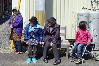 仮設住宅の道路沿いに設置された木製ベンチに、帽子やジャケットなど様々な服装をした4人の人物が座って、オスッサマの舞を見ている様子の写真
