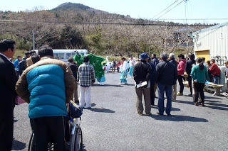 屋外の駐車場のような場所に人々が集まっており、獅子舞の被り物をしたオスッサマと装束姿の人が向かい合って舞を披露している様子を遠巻きに見ている写真