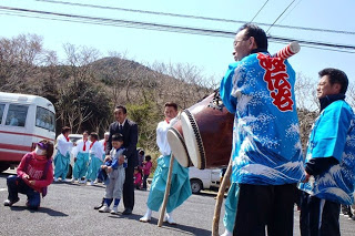 バスが停まっている駐車場で、波模様の青と白の法被を着た人や、白装束姿の人が太鼓を演奏している様子の写真
