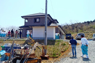 小高い山の麓に建てられた2階建ての新築住宅の周りに、白装束姿の人達が集まっている様子の写真