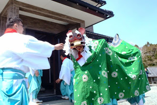 住宅の玄関の前に白装束姿の人たちが集まり、獅子舞の被り物をしたオスッサマ役の先頭の人が、白装束姿の男性からお酒の様な物を渡されている様子の写真
