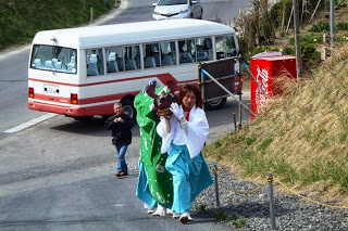 赤色のラインが入った白いバスをバックに、オスッサマ役の前に入っていた白装束姿の男性が、獅子舞の被り物を外して坂道を歩いている様子の写真