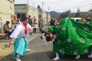 仮設住宅が建っている傍で、住宅に住んでいる人たちが見ているなか、獅子舞の被り物をしたオスッサマと、榊を持った装束姿の人が向かい合って舞を披露している様子の写真