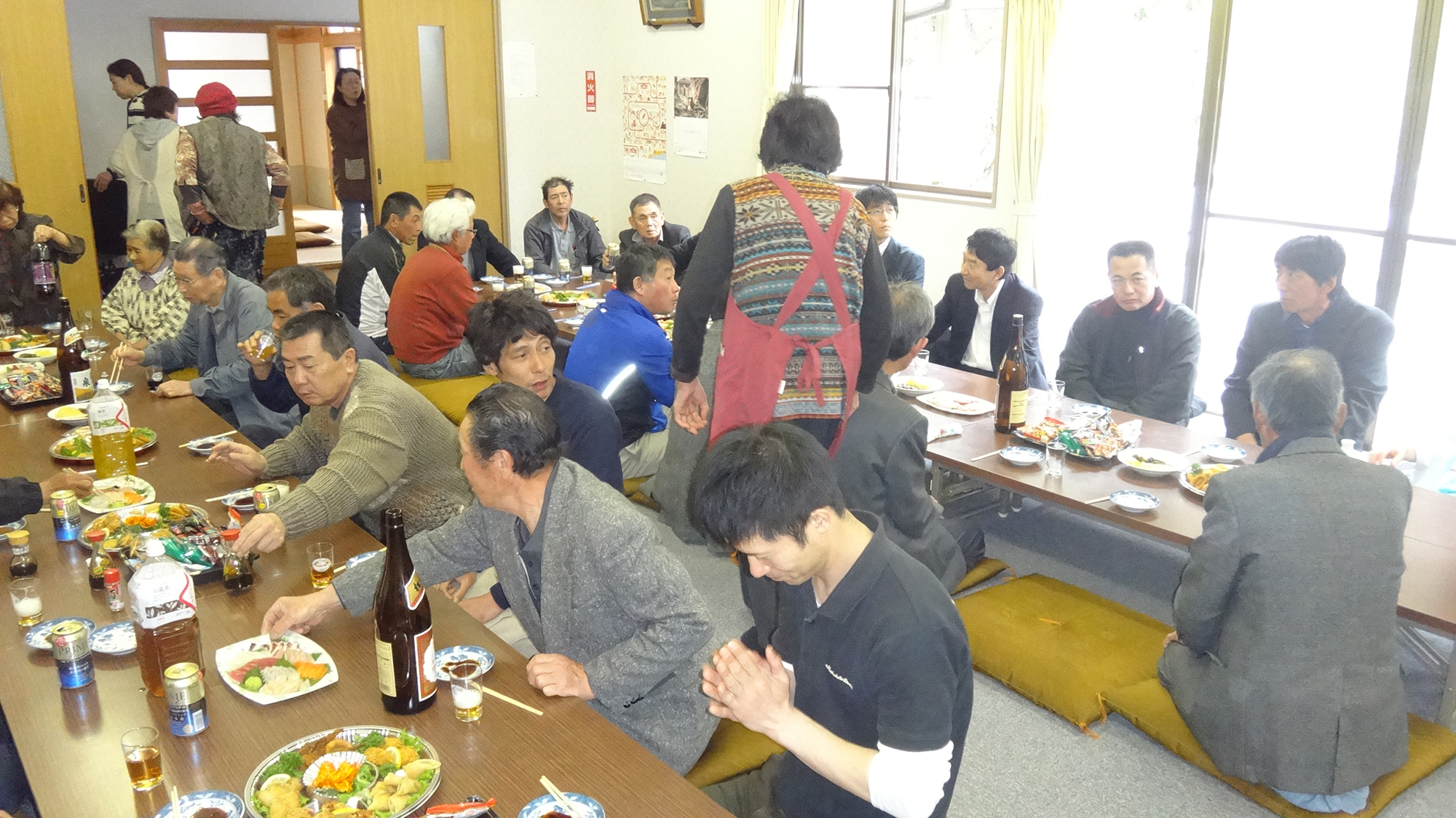 室内に2列設けられた長い座卓を囲み、たくさんの人々が料理と酒を楽しみながら交流している写真