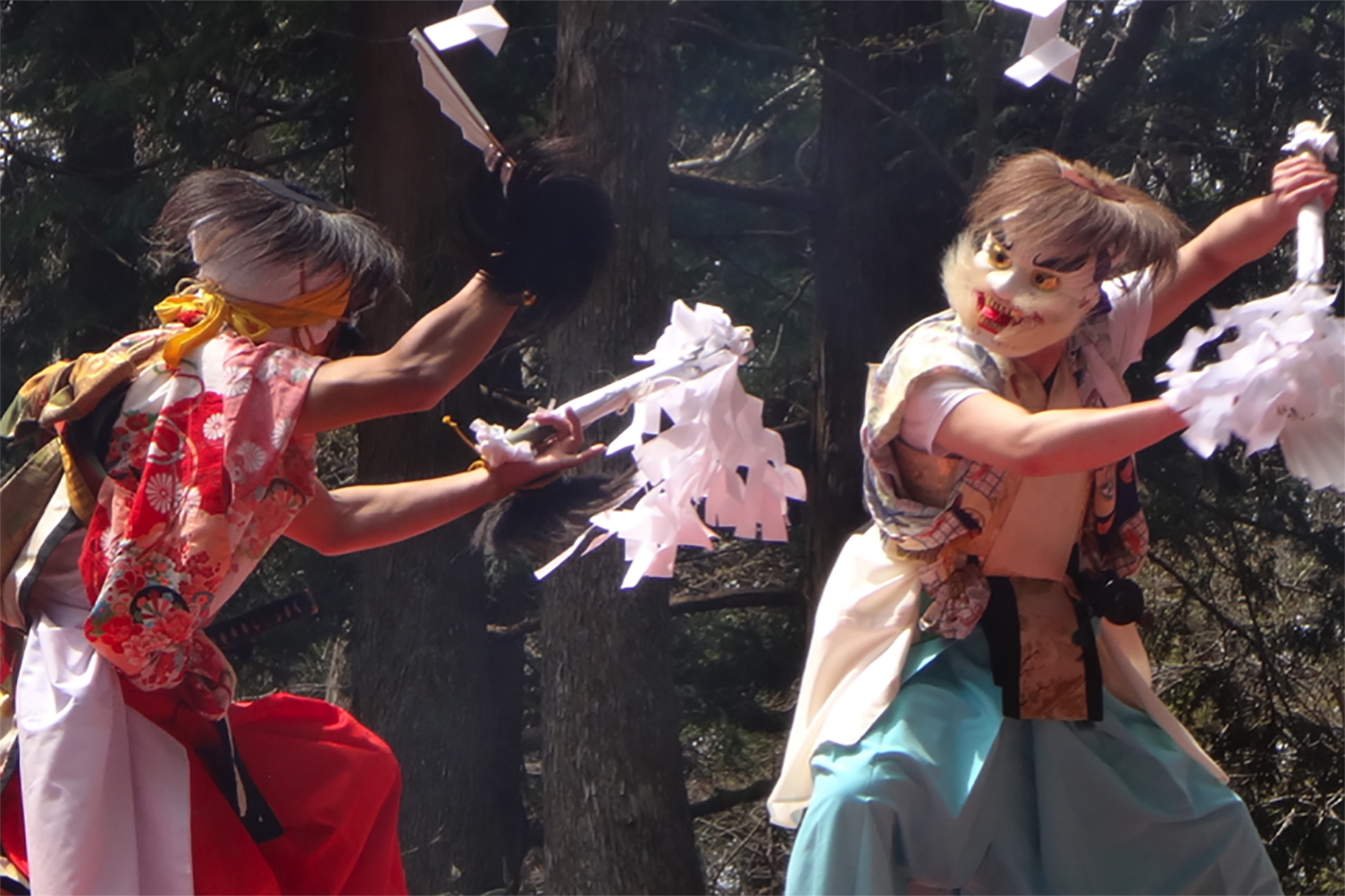 木立の中で面をつけた二人が剣を手に舞を披露している写真