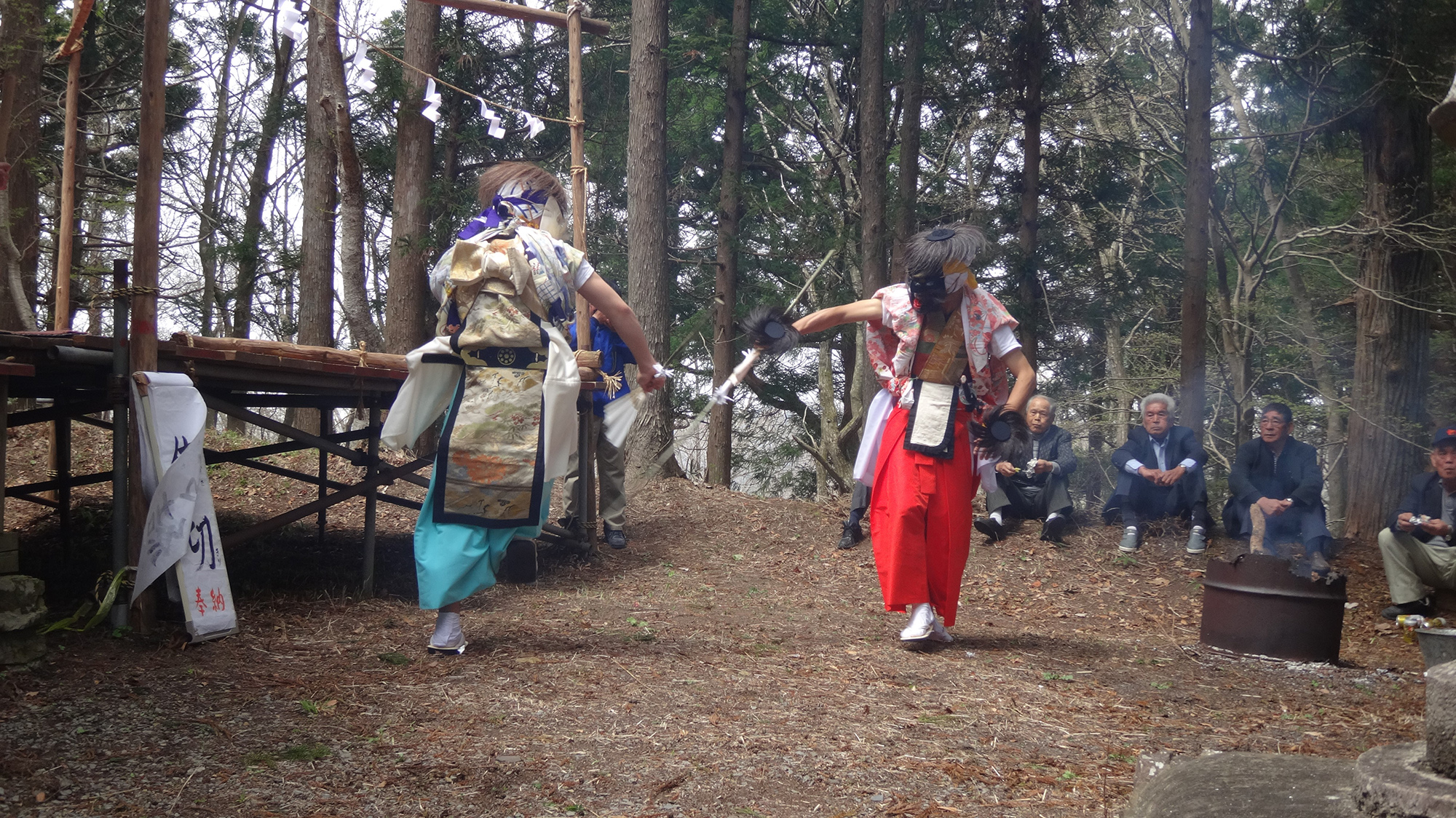 木立の中で面をつけた二人が向かい合い舞を披露し、周囲で人々が座って見守っている写真