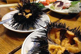 割られたウニの殻の中に黄色い身が詰まっており、刺身の盛り合わせと一緒に食卓に並べられている様子の写真