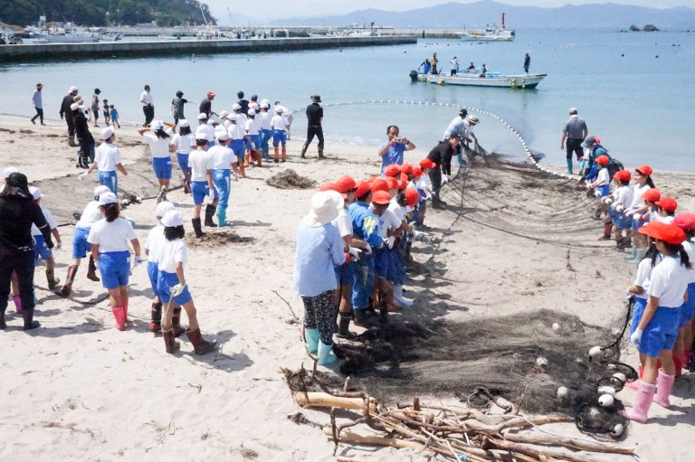 白と赤の帽子をかぶった小学生と大人たちが、浜辺で協力しながら大きな定置網を引っ張っている漁業体験学習の写真