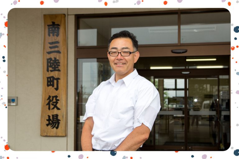 木製の看板に「南三陸町役場」と書かれた建物の前で、白いシャツを着た男性が笑顔で立っている様子をアップで撮影した写真