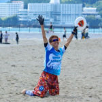 カラフルなシャツと短パンを着た佐藤慶治さんが、砂浜でフリスビーをキャッチしてひざまずき、両手を高く上げて喜んでいる写真