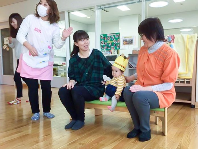 保育施設の室内で、ピンクのエプロン姿の保育士と別の女性が動いており、小さなベンチに座っている二人の女性が優しく見守っているなか、中央に手作りの紙で作られた王冠をかぶった幼児が座っているお誕生日会の様子の写真