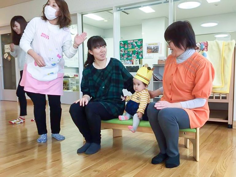 保育施設の室内で、ピンクのエプロン姿の保育士と別の女性が動いており、小さなベンチに座っている二人の女性が優しく見守っているなか、中央に手作りの紙で作られた王冠をかぶった幼児が座っているお誕生日会の様子の写真