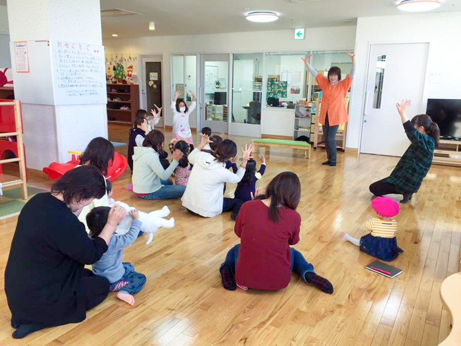 広い室内の前方で、オレンジの服を着た保育士が手を上げ体操のような動きを教えていて、同じように両手を上げた子どもとその保護者が輪になって座っている写真