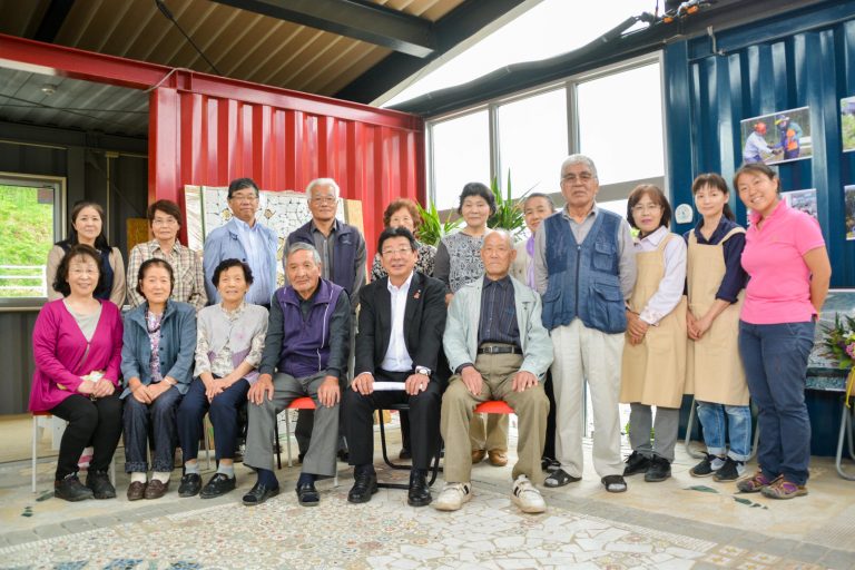 高齢者を中心とした地域の人々16人が赤と青のコンテナハウスの前で、前列に座った町長を中心に整列し、笑顔で写っている集合写真