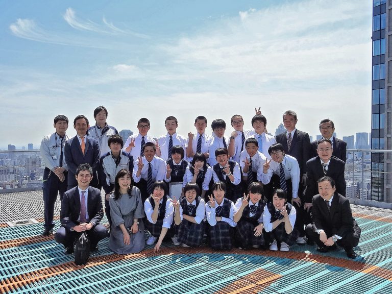 制服姿の高校生とスーツ姿の大人たち約25人が、ヘリポートのある高層ビルの屋上で、街並みを背景に笑顔でポーズをとって並んでいる写真
